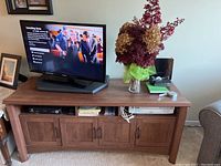 Wood TV cabinet holding a TV, vase with dried flowers, and decorative items on top. Four closed doors and three open storage compartments visible.