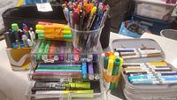 Photo of pen and marker assortment with some tools in clear acrylic organizer and others in zippered black compartment bag, showing various colors and brands