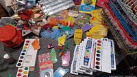 Overview photograph showing various art supplies including watercolor paint sets, magnetic pointing finger accessories, craft feathers, chalk packs, and storage containers.