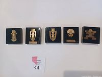 Five gold anthropomorphic pendants encased in individual black-backed acrylic blocks, shown front side.