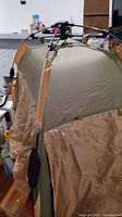 Photo of tent partially set up with beige and green fabric and metal poles with black connectors at top.