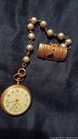 Full view of pocket watch attached to short pearl string with miniature souvenir pocketbook.
