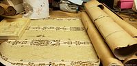 Seven antique wallpaper rolls and two border rolls rolled on a workbench with a beige base and brown floral geometric pattern.