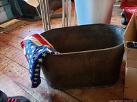 Photo displaying large antique copper tub on wood floor, with an American flag draped over edge, tub patinated and showing age wear.