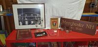 Framed black and white photo of Notre Dame Bridge with beer glasses, BMW champagne flutes, plaque, fireman book and wooden Manchester NH sign arranged on a red table.