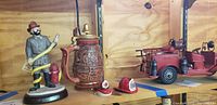 Photo showing Emmitt Kelley fireman figurine holding a hose around a hydrant, beer stein with detailed fire relief, two small fireman helmets, and a red toy fire truck on a wooden shelf.