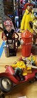 Photo showing four fireman-themed figurines including caricature fireman with extinguisher, fireman on fire truck toy, two fireman figurines in colorful outfits, and a red fire hydrant wall-hanging.