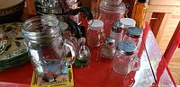 Several glass mason jars of varying sizes on a shelf with metal lids including shaker tops and jars with handles, a black-handled ladle, and a mason jar grip.