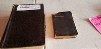 Two tiny antique books, one large and rectangular with a black cover, and a smaller black leather prayer book titled 'Treasury Prayer'. Both show signs of wear and age.