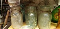 Three vintage Mason jars varying in size, with glass lids and wire bail closures on wooden shelf.