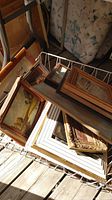 Bin of assorted framed art paintings stacked inside a basket on wooden deck, showing range of frame styles and sizes.