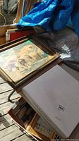 Photo shows a framed art print with rural scene including an ox and family near haystacks, a white box for the art book, with some frames stacked beneath covered by tarps.