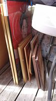 Multiple framed art pieces stacked and leaning against a wall outdoors, showing edges and frame details.