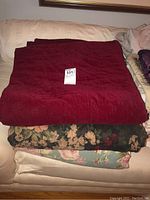 Three folded blankets stacked on a couch: solid dark red quilted velvet-like blanket on top, dark floral patterned blanket in the middle, and teal floral patterned blanket at the bottom.