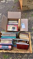 Photo shows basket and cardboard box filled with assorted books, laid outdoors on ground with grass and dirt visible.