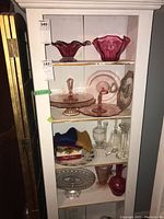 Front view of white bookshelf filled with pink glass bowls and cake stands on top shelf, clear glass decanters and candlesticks, fabric pieces, and decorative plates on middle shelves, blue and white ceramic and silver accented plates on bottom shelf.