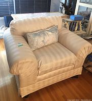 Front and angled view of beige striped upholstered feather filled armchair with rolled arms, skirted base, removable cushions, and decorative pillow