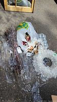 Photo of white artificial wreath, bundle of decorative branches, bag containing Christmas decor, and white string lights all laid on pavement.