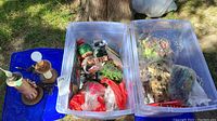 Two plastic bins packed with Christmas decorations and crafting supplies and two wooden base lamps on blue lid outdoors on grass and ground.