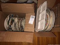 Two open cardboard boxes filled with assorted plates, platters, and bowls. Plates have various decorative and floral designs. White ceramic platters with wavy edges seen on top.