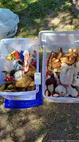 Two plastic bins filled with assorted dolls and bears outdoors on grass.