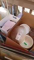 Box containing a white ceramic serving dish with lid, a large white bowl, and a clear ribbed glass ceiling shade dome, with some folded linens.