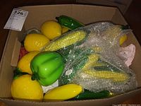 Box filled with various glass fruits and vegetables, including peppers, lemons, corn, and bananas, with bubble wrap for protection.
