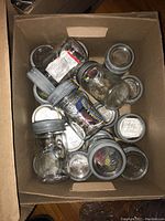 Top-down view of 18 glass mason jars in a cardboard box, showing the jars with silver metal screw-on lids and embossed designs on the glass.