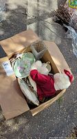 Cardboard box containing white teddy bear dressed in red and several plates along with a rectangular glass vase, all arranged loosely.