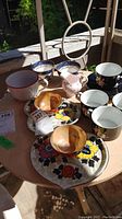 Ten assorted vintage teacups and saucers displayed on a table outdoors including floral, blue and white, and pig-shaped pieces.