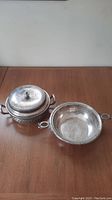 Two silver serving dishes on wooden table, one with lid and ornate floral detailing, the other smooth bowl shape.