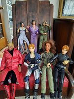 Seven action figures displayed on wooden chair: front row includes Dylan Sanders in red trenchcoat, metallic silver-pants figure, green-jacketed figure and black jumpsuit figure; back row includes purple tracksuit figure, gray dress female figure and camo uniform figure.