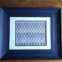 Full view of the framed tapestry on a wooden surface showcasing the dark wooden frame and white and brown double matting around the textile artwork.