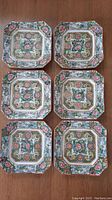 Six hand-painted square porcelain plates arranged in two columns of three on a wooden surface.