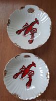Two white scalloped bowls with bright red lobsters and gold edge trim shown together