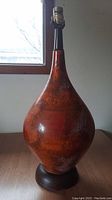 Full view of the mid-century modern table lamp showing the bulbous ceramic body with reddish-brown mottled glaze, wooden base, and lamp socket on top.