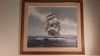 Framed oil painting hung on wall showing a sailing ship on the ocean under a cloudy sky, signed Alex, wooden frame with beige mat.