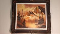 Original framed oil painting showing a mountain landscape with trees and reflection, surrounded by an ornately carved wooden frame with visible corner damage.