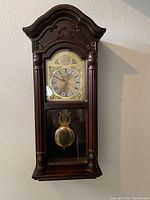 Wall-mounted Bedford chime clock with wooden ornate casing, circular Roman numeral clock face, and brass pendulum.