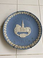 Top-down photo of blue jasperware plate with white relief image of a church, the word 'CHRISTMAS' and the year '1983' with a leaf border.