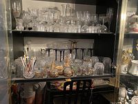 Photo of a black cabinet filled with various crystal glassware on two shelves, including glasses, decanters, tumblers, shot glasses, and glass figurines.