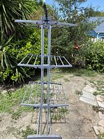 Three-tier gray plastic clothes drying rack shown outdoors in garden setting. Structure has multiple horizontal rods on three levels for hanging clothes. Rack has wheels and plastic frame tubes. Rack is standing upright, partially folded.