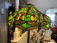 Green Tiffany-style stained glass floor lamp shade featuring dragonfly designs with red, green, and amber glass sections. Shade is scalloped along the edges.