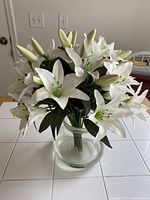 Artificial white lily floral arrangement in a clear glass bowl vase on a white surface against a neutral background.