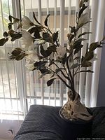 Imitation lily plant with white flowers and green leaves placed on a dark cushion in front of vertical blinds.