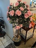 Artificial rose plant about 50 inches tall with many pink flowers and green leaves, in black pot, shown in room corner.