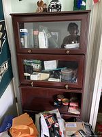 Photo shows a tall wooden office bookcase with three glass front opening sections. The shelves contain miscellaneous boxed items and papers.