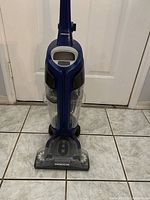 Oreck SurfaceScrub upright vacuum cleaner standing on tiled floor showing front, dirt canister, and cleaning head with Oreck branding.