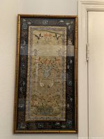 Frontal image of framed oriental silk embroidery textile hanging on a wall, showing detailed floral and bird designs with a decorative border and gold frame.