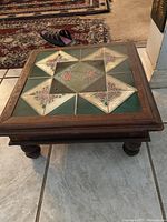 Square wooden display stand with a patterned tiled top in green, cream and pink floral design, thick wooden frame and round legs.